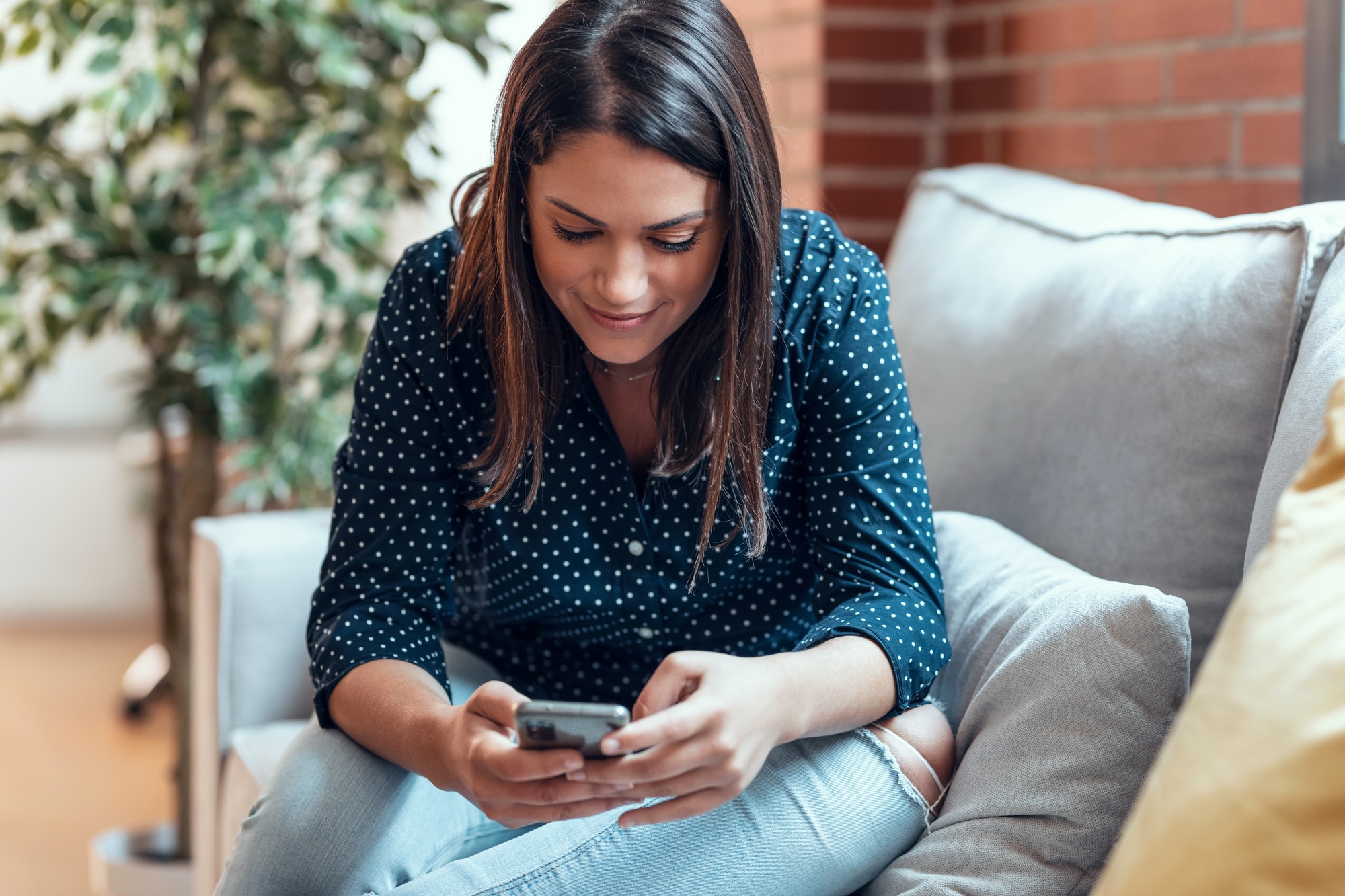 Beautiful woman sending messages with her mobile phone while sitting on a couch at home.